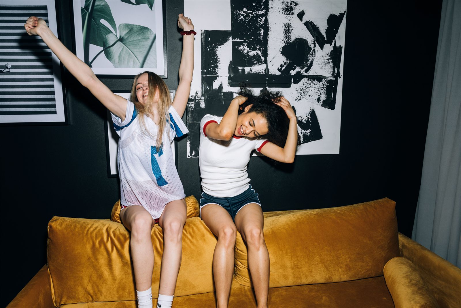 Zwei Frauen tanzen auf einem senfgelben Sofa vor dunkler Wand mit Grafiken